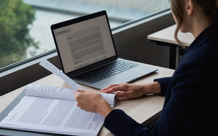 A woman at a desk reading a printed policy document beside an open laptop
